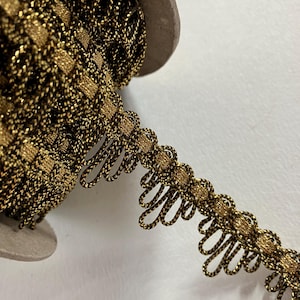 May include: A spool of decorative trim featuring a gold and black woven pattern. The trim has a scalloped edge with looped details. The spool is made of cardboard. The trim is suitable for sewing, crafting, and embellishing projects.