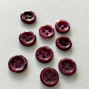 May include: A collection of nine round, burgundy buttons with four holes each. The buttons have a pearlescent sheen, reflecting light with a subtle iridescence. These sewing notions are ideal for adding a decorative touch to garments or crafts.