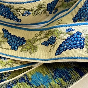 May include: Close-up of decorative ribbon featuring a repeating pattern of blue grapes and green leaves on a cream background. The ribbon has a blue border and a fringed edge with blue and green threads. Ideal for crafting and embellishments.