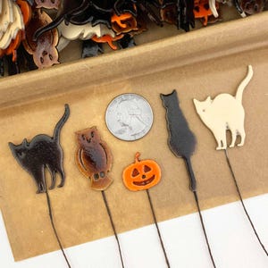 May include: Halloween-themed decorative picks featuring a black cat, owl, pumpkin, and a white cat. The picks are on thin wire stems. A U.S. quarter is included for size reference. The items are set on a brown surface.