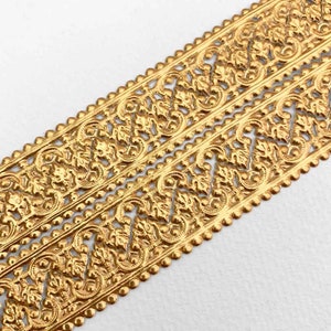 May include: Close-up of two parallel strips of gold-colored decorative trim. The trim features an ornate, repeating pattern of leaves and swirls, with a beaded edge. The design is detailed and intricate, suggesting a vintage or antique aesthetic. The background is white.