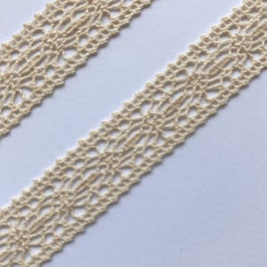 May include: Two strips of cream-colored lace with an openwork design. The lace has a delicate, intricate pattern with repeating floral or geometric shapes. The lace is likely made of cotton or a similar natural fiber.