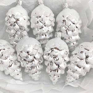May include: A collection of silver pinecone-shaped Christmas ornaments. Each ornament is covered in a dusting of white snow, creating a festive and wintery appearance. The ornaments are arranged in a white box, ready for display.