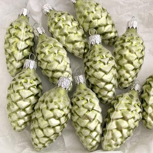 May include: A collection of 12 green glass ornaments shaped like pine cones. The ornaments have a frosted finish and are decorated with a silver cap.