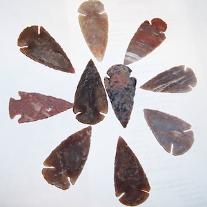 May include: A collection of ten stone arrowheads arranged in a circular pattern. The arrowheads vary in color, including shades of brown, red, and tan, with unique patterns and textures. Each arrowhead has a distinct notched base.