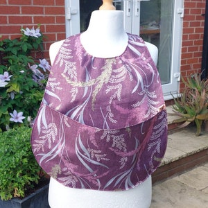 May include: A purple bib with a pocket, featuring a botanical pattern of ferns and leaves in shades of lavender, white, and green. The bib is displayed on a white mannequin head and torso, showcasing its shape and design.