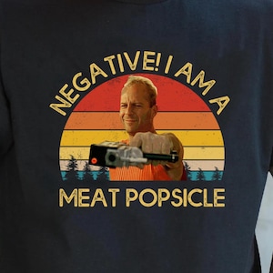 May include: Black t-shirt with a retro graphic featuring a man holding a weapon, set against a sunset backdrop. The text reads "NEGATIVE! I AM A MEAT POPSICLE" in a vintage font. A humorous and pop culture-themed apparel item.
