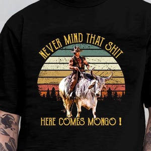May include: Black t-shirt featuring a graphic of a cowboy riding a bull in front of a sunset. The text reads "NEVER MIND THAT SHIT" above and "HERE COMES MONGO!" below the image. The design has a vintage, retro style.