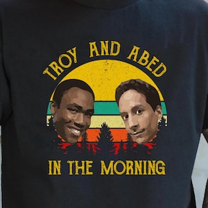 Könnte beinhalten: Schwarzes T-Shirt mit Retro-Design. Der Text lautet "TROY AND ABED IN THE MORNING". Die Grafik zeigt zwei Porträts vor einem gelb-grün gestreiften Sonnenuntergang mit einer Waldsilhouette.