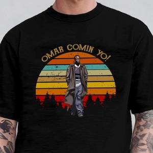 May include: Black t-shirt with the text "OMAR COMIN YO!" above a graphic of a man in a long coat. The graphic is set against a sunset design with trees.