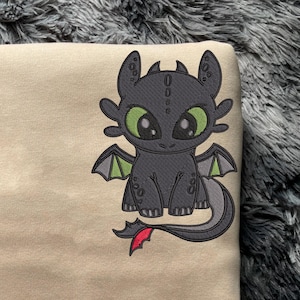 May include: A beige sweatshirt with a black and green cartoon dragon embroidered on the front. The dragon has large eyes and a red tail.