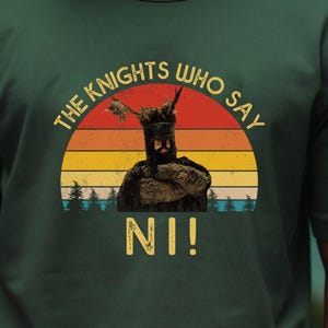 Peut inclure: T-shirt vert foncé avec un motif de chevalier devant un coucher de soleil. Le texte "THE KNIGHTS WHO SAY NI!" est disposé en arc de cercle au-dessus du chevalier, avec "NI!" en dessous. Le t-shirt est de style col rond.