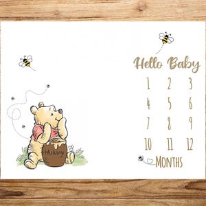 May include: White baby milestone blanket featuring Winnie the Pooh with a honey pot and bees. The blanket has the text "Hello Baby" and numbers 1-12 for monthly photos. A cute baby shower gift.