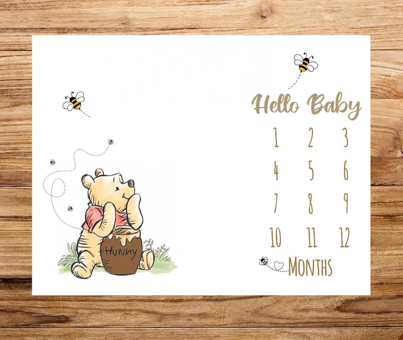 May include: White baby milestone blanket featuring Winnie the Pooh with a honey pot and bees. The blanket has the text "Hello Baby" and numbers 1-12 for monthly photos. A cute baby shower gift.