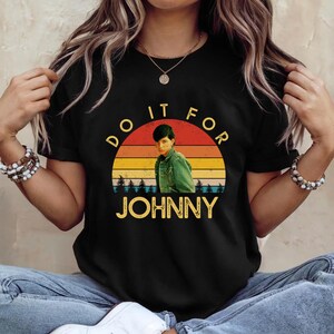 May include: Black t-shirt featuring a retro sunset design with the text "DO IT FOR JOHNNY." The graphic includes an image of a person in a green jacket. The word "JOHNNY" is in a yellow font.