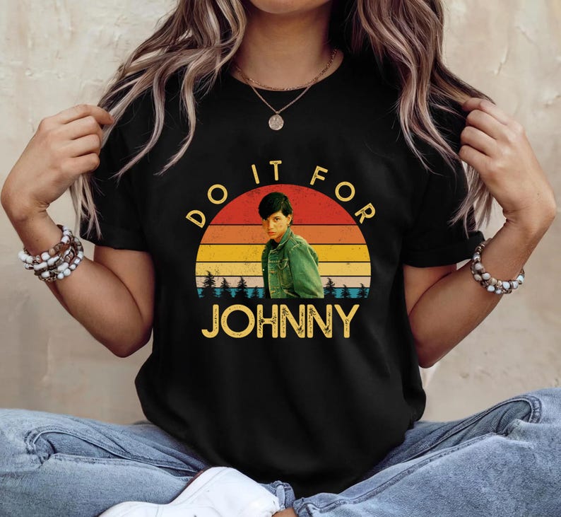 May include: Black t-shirt featuring a retro sunset design with the text "DO IT FOR JOHNNY." The graphic includes an image of a person in a green jacket. The word "JOHNNY" is in a yellow font.