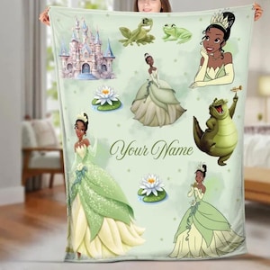 May include: A light green blanket featuring illustrations of a princess, a castle, frogs, and water lilies. The blanket has the text "Your Name" in a script font. The princess is wearing a green and yellow gown.