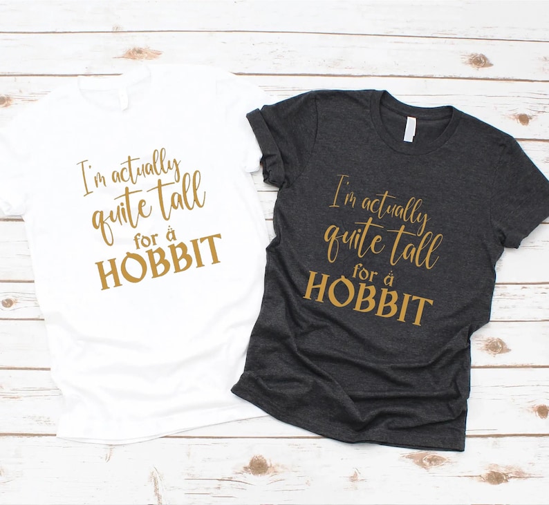 May include: Two t-shirts, one white and one dark gray, with the text "I'm actually quite tall for a HOBBIT" in gold lettering. The shirts are short-sleeved and appear to be made of a soft material.