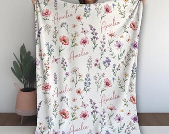 May include: A white throw blanket featuring a repeating pattern of watercolor-style wildflowers, small butterflies, and the name Amelia printed in a cursive script. The floral design includes various stems in shades of pink, purple, blue, and green.