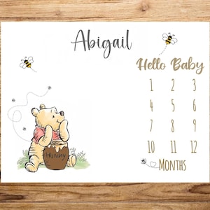 May include: White milestone blanket featuring a Winnie the Pooh illustration with the name "Abigail" in script. The blanket includes the text "Hello Baby" and numbers 1-12 for monthly photos. Bees and a honey pot are also featured.