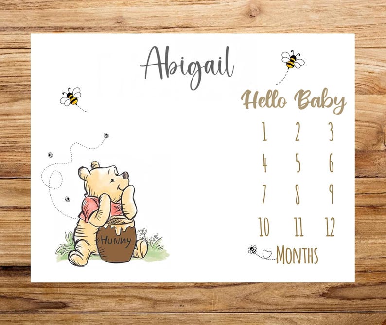 May include: White milestone blanket featuring a Winnie the Pooh illustration with the name "Abigail" in script. The blanket includes the text "Hello Baby" and numbers 1-12 for monthly photos. Bees and a honey pot are also featured.