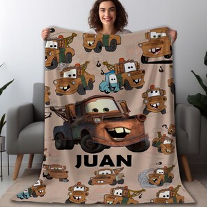 Puede incluir: Una manta de forro polar de color beige con un patr&oacute;n repetido de camiones de remolque de dibujos animados en varios colores. La manta tiene el nombre "JUAN" en letras negras.