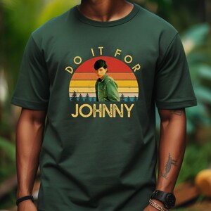 May include: Dark green t-shirt featuring a retro graphic with the text "DO IT FOR JOHNNY". The graphic includes a portrait of a person in a green jacket set against a sunset design with the name "JOHNNY" in gold letters.