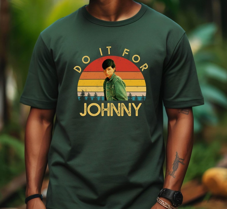 May include: Dark green t-shirt featuring a retro graphic with the text "DO IT FOR JOHNNY". The graphic includes a portrait of a person in a green jacket set against a sunset design with the name "JOHNNY" in gold letters.