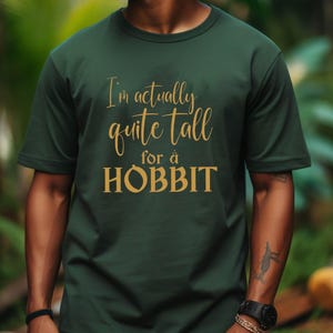 May include: A dark green t-shirt with the text "I'm actually quite tall for a HOBBIT" in gold lettering. The shirt is a crew neck style and appears to be made of a soft material. The image is a medium shot of the shirt on a person.