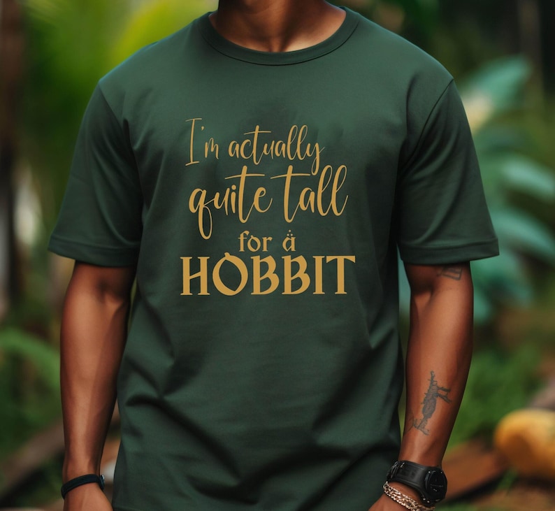 May include: A dark green t-shirt with the text "I'm actually quite tall for a HOBBIT" in gold lettering. The shirt is a crew neck style and appears to be made of a soft material. The image is a medium shot of the shirt on a person.