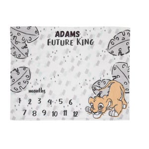 May include: White baby milestone blanket with a cartoon lion cub illustration. The blanket features the text "ADAMS FUTURE KING" and numbers for tracking baby's age in months. Decorative cheese-shaped elements are also present.