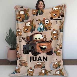 Puede incluir: Una manta de franela marr&oacute;n y naranja con tem&aacute;tica de Cars. La manta presenta un patr&oacute;n repetitivo de Mater, Rayo McQueen y otros personajes de la pel&iacute;cula. La manta est&aacute; personalizada con el nombre "JUAN" en letras negras.