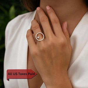 May include: A silver ring with a circular design and a small, clear gemstone set inside. The ring is worn on a finger, highlighting its minimalist and modern style. The ring's design is simple yet elegant, suitable for everyday wear.