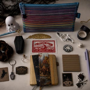 Könnte beinhalten: Eine Sammlung von Schreibwaren und Dekorationsartikeln, darunter ein gestreiftes Federmäppchen, ein goldenes Notizbuch mit Lesezeichen, eine schwarze sargförmige Schachtel, eine Geisterfigur, ein Mond- und Sonnenanhänger, Washi-Tape und kleine Anhänger.