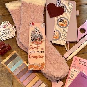 May include: A collection of reading accessories and cozy items. Includes a pair of pink fuzzy socks, a bookmark with a skeleton design, a heart-shaped felt embellishment, sticky notes, a notebook, and a sticker that says "Spiritually I'm at The Scholastic Book Fair."