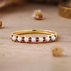 May include: A gold band ring featuring an alternating pattern of small white pearls and tiny red rubies set along the front half of the band. The ring has a polished finish and a thin, delicate profile, showcasing a symmetrical arrangement of gemstones.