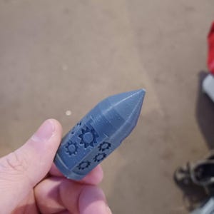 May include: A gray 3D printed rocket-shaped object with a pointed tip and a design of gears on the side.