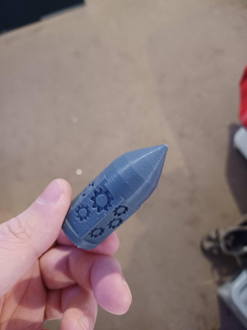 May include: A gray 3D printed rocket-shaped object with a pointed tip and a design of gears on the side.