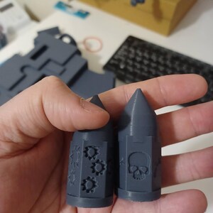 May include: Two gray 3D printed bullet-shaped dice with different designs. One die has a skull and crossbones design, the other has a gear design.