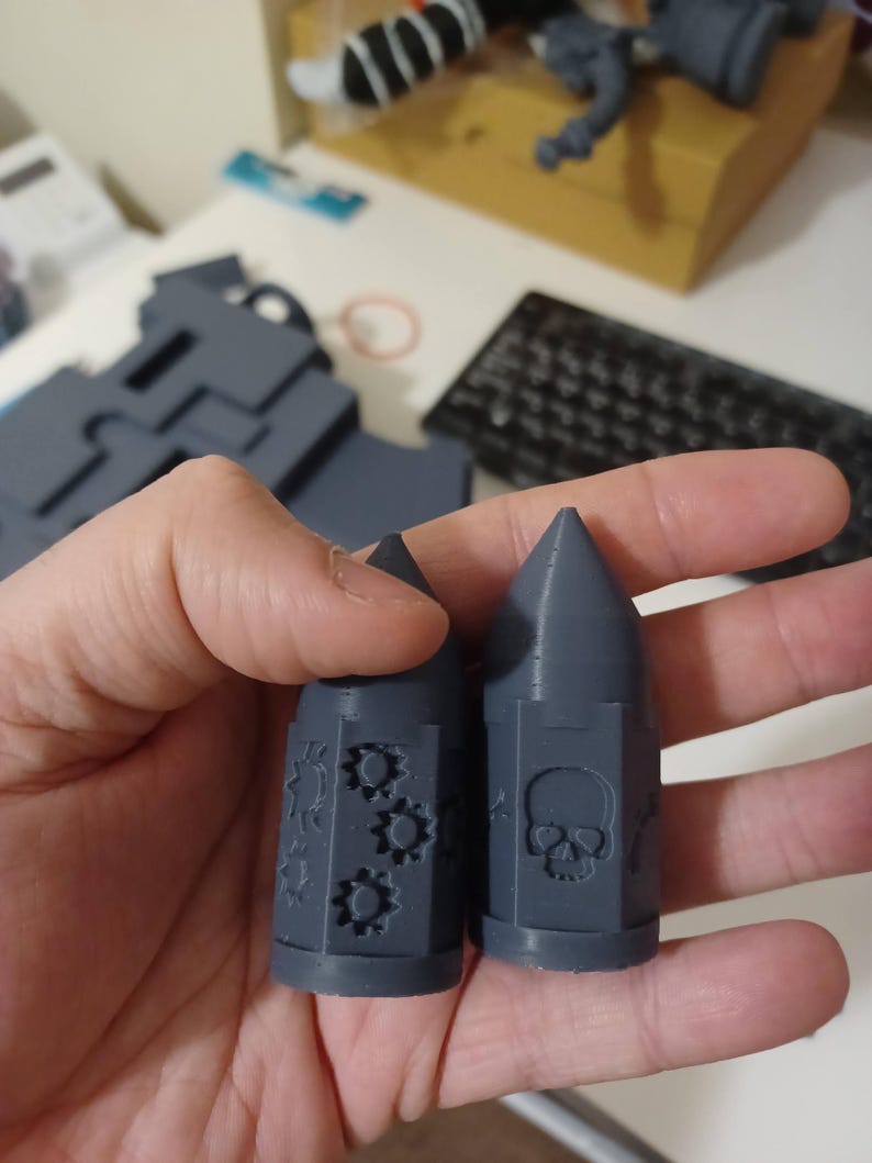 May include: Two gray 3D printed bullet-shaped dice with different designs. One die has a skull and crossbones design, the other has a gear design.