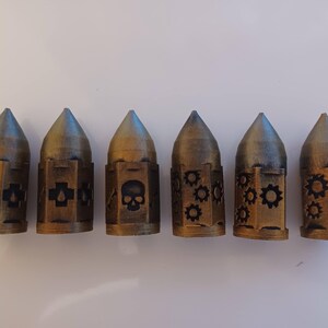 May include: Six small, gold-colored metal containers with different designs. Each container has a pointed top and a cylindrical body. The designs include a skull, a cross, a bone, and gears.