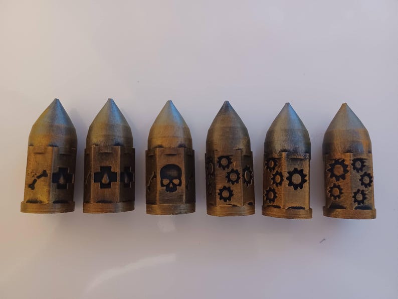 May include: Six small, gold-colored metal containers with different designs. Each container has a pointed top and a cylindrical body. The designs include a skull, a cross, a bone, and gears.