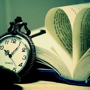 May include: A dark-toned pocket watch with a white face, black numerals, and ornate hands rests beside an open book. The pages of the book are folded to form a heart shape, with visible printed text on the paper. The scene has a cool, greenish tint.