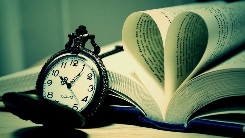 May include: A dark-toned pocket watch with a white face, black numerals, and ornate hands rests beside an open book. The pages of the book are folded to form a heart shape, with visible printed text on the paper. The scene has a cool, greenish tint.