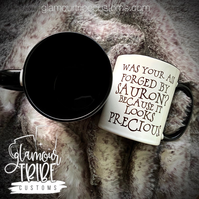 May include: Two ceramic mugs on a faux fur surface. One mug is black, the other is white with black text that reads "WAS YOUR ASS FORGED BY SAURON? BECAUSE IT LOOKS PRECIOUS." The Glamour Tribe Customs logo is visible.