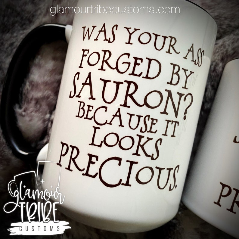 May include: White ceramic mug with a black handle and rim, featuring the text "WAS YOUR ASS FORGED BY SAURON? BECAUSE IT LOOKS PRECIOUS" in dark brown lettering. The mug has the Glamour Tribe Customs logo.