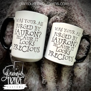 May include: Two white ceramic mugs with black handles and rims. The mugs feature the text "WAS YOUR ASS FORGED BY SAURON? BECAUSE IT LOOKS PRECIOUS" in black lettering. The Glamour Tribe Customs logo is visible.