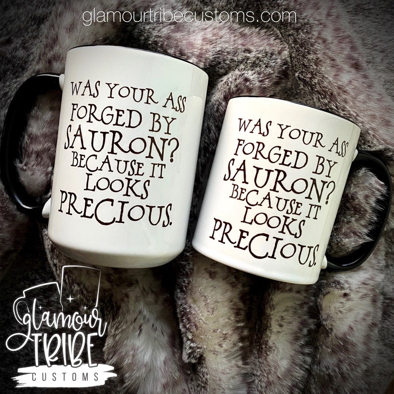 May include: Two white ceramic mugs with black handles and rims. The mugs feature the text "WAS YOUR ASS FORGED BY SAURON? BECAUSE IT LOOKS PRECIOUS" in black lettering. The Glamour Tribe Customs logo is visible.
