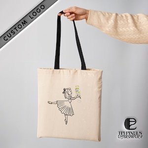 以下が含まれることがあります： 黒い持ち手付きのベージュのキャンバス地のトートバッグ。バッグには、くるみ割り人形を持ったバレリーナの刺繍が施されています。バッグの右下には「TRIPIXELS EMBROIDERY」の文字が印刷されています。