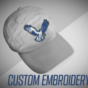 May include: A gray baseball cap with a blue embroidered hawk design. The cap has a curved brim and a traditional six-panel construction. The text "CUSTOM EMBROIDERY" is printed in blue at the bottom of the image.